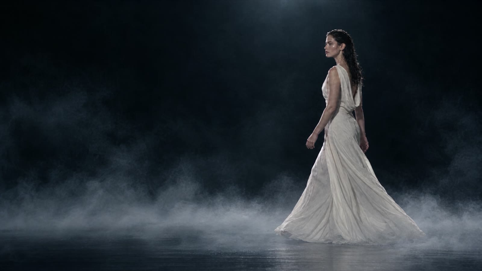 A model in a long white gown walking through fog on a dark stage